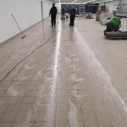Czyszczenie podłogi w sklepie za pomocą specjalistycznej maszyny, widoczne ślady wody i mydlin na kafelkach, pracownicy w tle.