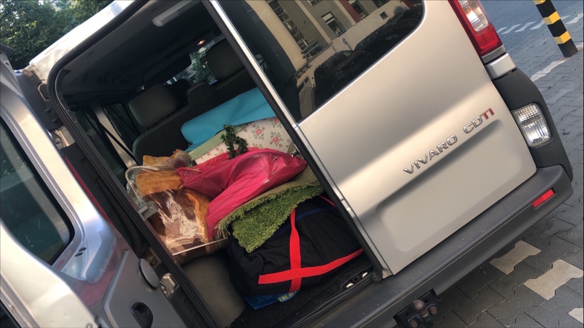 Otwarty bagażnik srebrnego vana Vivaro wypełniony spakowanymi rzeczami osobistymi, widoczne poduszki, koce i torby, przygotowany do transportu.