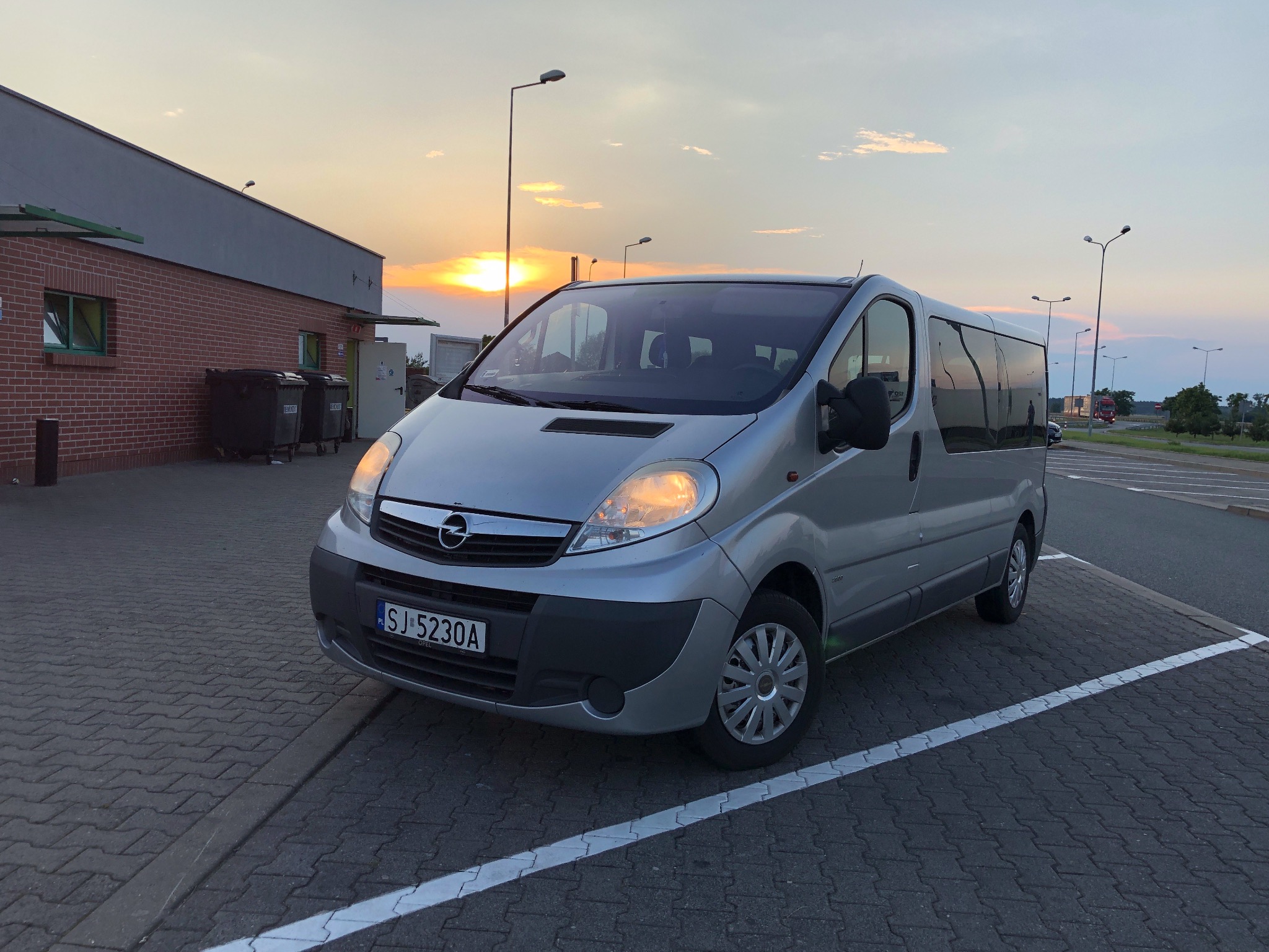 Srebrny Opel Vivaro zaparkowany na parkingu z kostki brukowej, w tle budynek z cegły i zachodzące słońce.