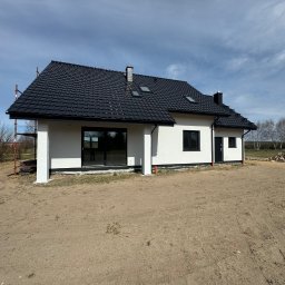 S-bud Sylwester Bonk - Nowoczesny dom jednorodzinny z czarnym dachem, białą elewacją i dużymi oknami, widoczny z zewnątrz, na działce z piaskiem. Częściowo widoczne rusztowanie.