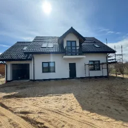 Nowoczesny dom z czarnym dachem i białymi ścianami w słoneczny dzień. Widoczne rusztowania, otwarty garaż i składowane drewno obok.