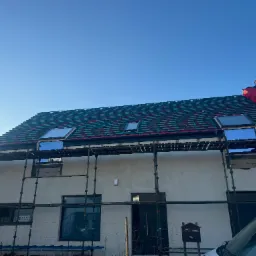 Dom w trakcie krycia dachu z oknami dachowymi, widoczne rusztowania i folia dachowa. Częściowo otynkowana elewacja, błękitne niebo w tle.