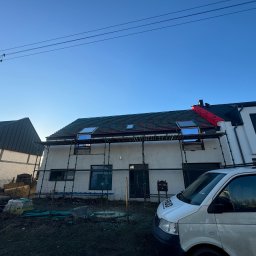 S-bud Sylwester Bonk - Dom w trakcie krycia dachu z oknami dachowymi, widoczne rusztowania i fragment firmowego busa. Częściowo położona dachówka, błękitne niebo w tle.