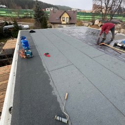 S-bud Sylwester Bonk - Prace dekarskie na płaskim dachu: dekarz w czerwonej koszuli układa papę termozgrzewalną. Widoczny sprzęt, butla z gazem i narzędzia dekarskie. W tle zabudowa jednorodzinna.