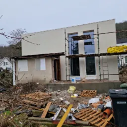 Dom w trakcie ocieplania elewacji, widoczne rusztowania, śmieci i materiały budowlane. Fragment budynku z nową elewacją. Tło: zalesione wzgórze.