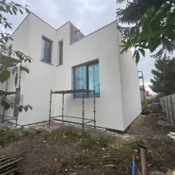 Biała elewacja nowoczesnego domu w trakcie wykańczania, okna zabezpieczone folią, widoczne rusztowania i teren budowy. Minimalistyczna architektura.