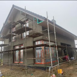 S-bud Sylwester Bonk - Dom w trakcie nakładania szarego tynku elewacyjnego, z rusztowaniami i oknami zabezpieczonymi folią. Widoczny fragment dachu i otoczenie placu budowy.