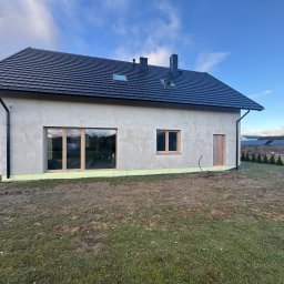 S-bud Sylwester Bonk - Niedokończony dom jednorodzinny z ciemną dachówką, otynkowany, z widocznymi oknami i drzwiami. W tle błękitne niebo i fragment ogrodu z tujami.