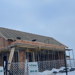 S-bud Sylwester Bonk - Budowa domu z czerwonej cegły w zimowej scenerii. Widoczne drewniane elementy konstrukcji dachu i rusztowania. Na pierwszym planie ogrodzenie z siatki i śnieg.