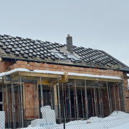 S-bud Sylwester Bonk - Budowa domu w zimowej scenerii: częściowo pokryty dach czarną dachówką, ceglane ściany w rusztowaniach, całość otulona śniegiem. Widoczny komin i drewniana konstrukcja dachu.