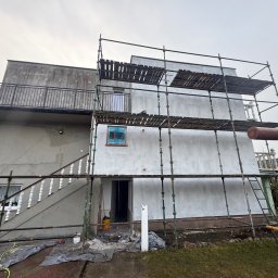 S-bud Sylwester Bonk - Dwupiętrowy dom w trakcie prac elewacyjnych, otoczony rusztowaniem. Ściany pokryte szarą warstwą, okno zabezpieczone folią. Widoczne schody zewnętrzne i balkon.