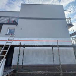 S-bud Sylwester Bonk - Szara elewacja domu w trakcie prac wykończeniowych. Widoczne rusztowania, drabina i fragmenty niezakończonej powierzchni. Na górze balkon z czarną balustradą.