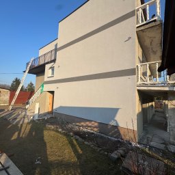 S-bud Sylwester Bonk - Dwukondygnacyjny dom z szarą elewacją i balkonami, z drabiną opartą o ścianę. Widoczne detale wykończenia i otoczenie domu w słoneczny dzień.