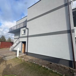 S-bud Sylwester Bonk - Dwukondygnacyjny dom z szarą elewacją, balkonem z czarną balustradą i schodami z białą balustradą. Widoczny fragment trawnika i rynna.