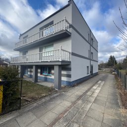 S-bud Sylwester Bonk - Dwupiętrowy dom z szarą elewacją i białymi balkonami, widoczny z perspektywy chodnika. Okna zabezpieczone folią. Na elewacji widoczne szare pasy.