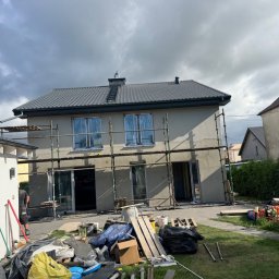 S-bud Sylwester Bonk - Dwupiętrowy dom jednorodzinny z ciemnoszarym dachem z dachówki, rusztowaniem na elewacji i oknami osłoniętymi niebieską folią. Na pierwszym planie materiały i odpady budowlane.
