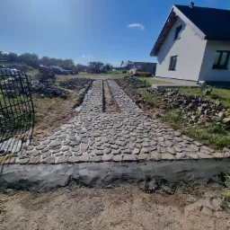 Budowa podjazdu z kamienia polnego z centralnym odpływem liniowym, widoczny etap prac ziemnych i fundament z betonu, w tle dom jednorodzinny.