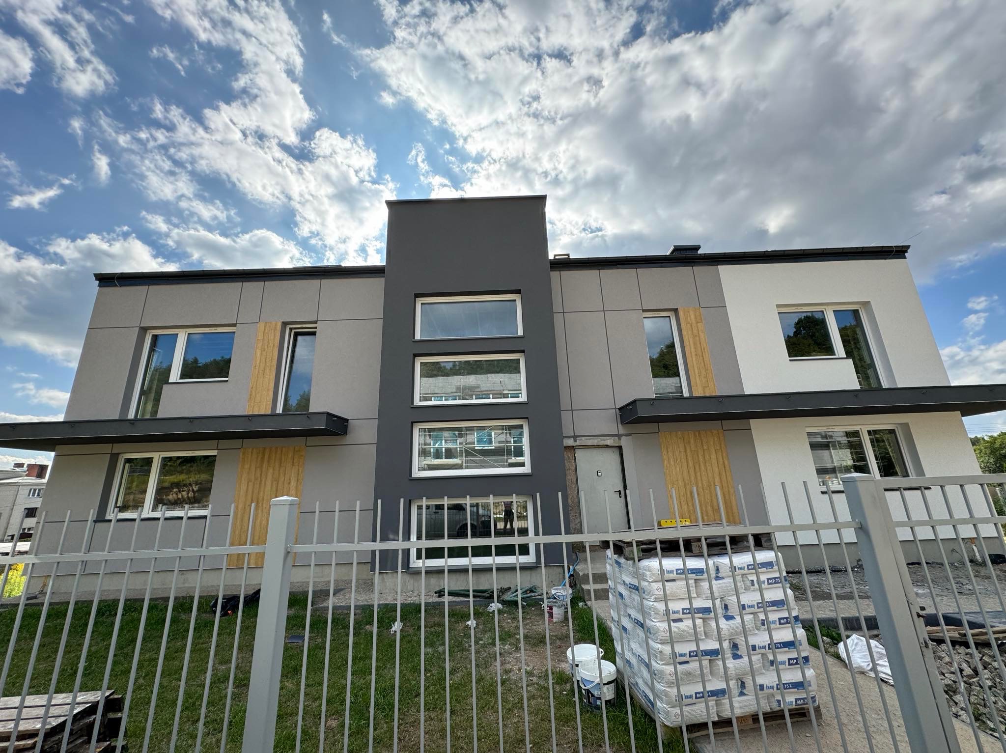Nowoczesny dwukondygnacyjny dom w trakcie wykańczania elewacji, widoczny przez metalowe ogrodzenie, z paletą worków z zaprawą i pojemnikami na farbę obok wejścia.