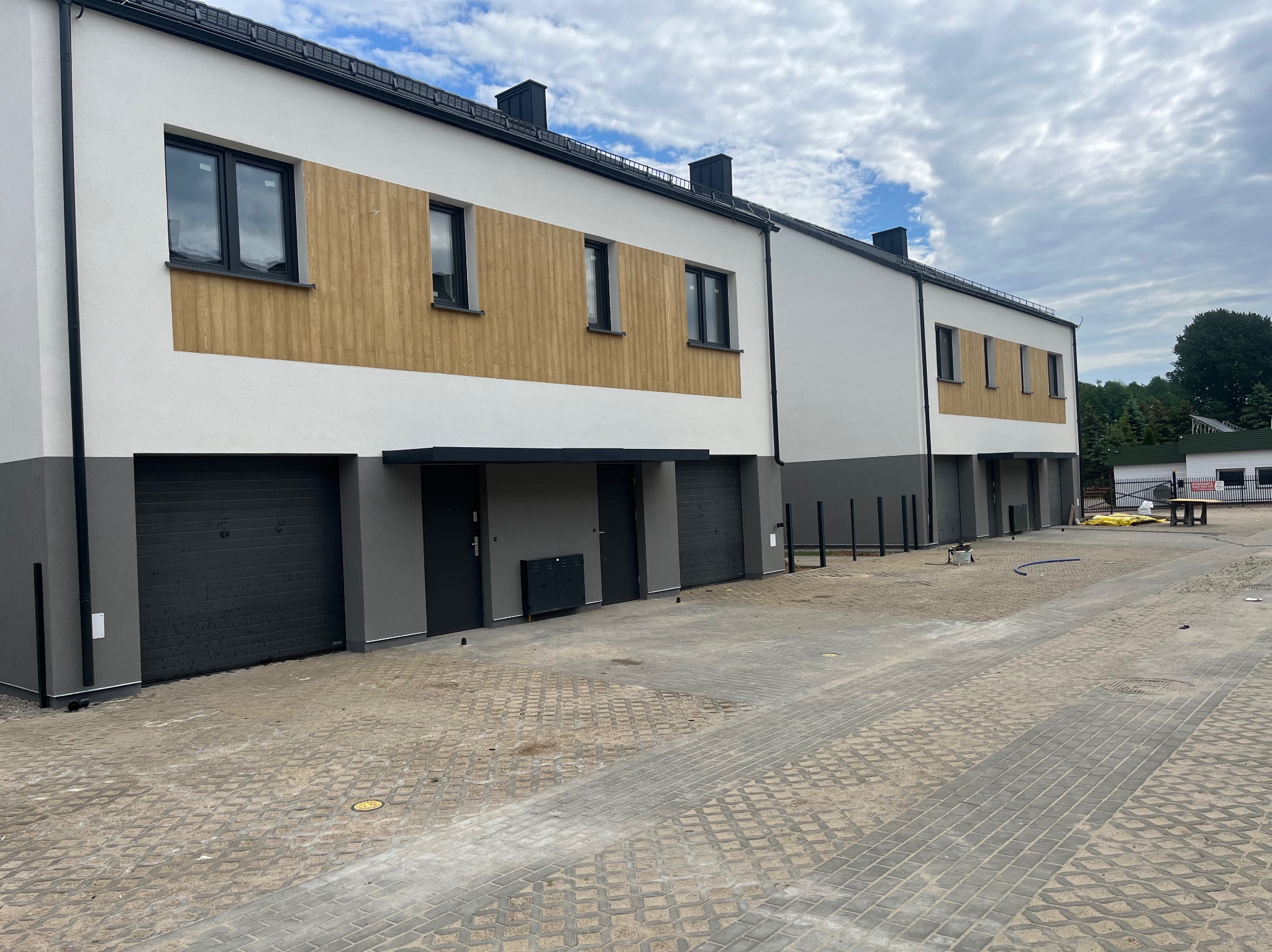Szereg nowo wybudowanych domów szeregowych z elewacją w kolorach bieli i szarości, akcenty z paneli drewnopodobnych, czarne okna i bramy garażowe, kostka brukowa na podjeździe.