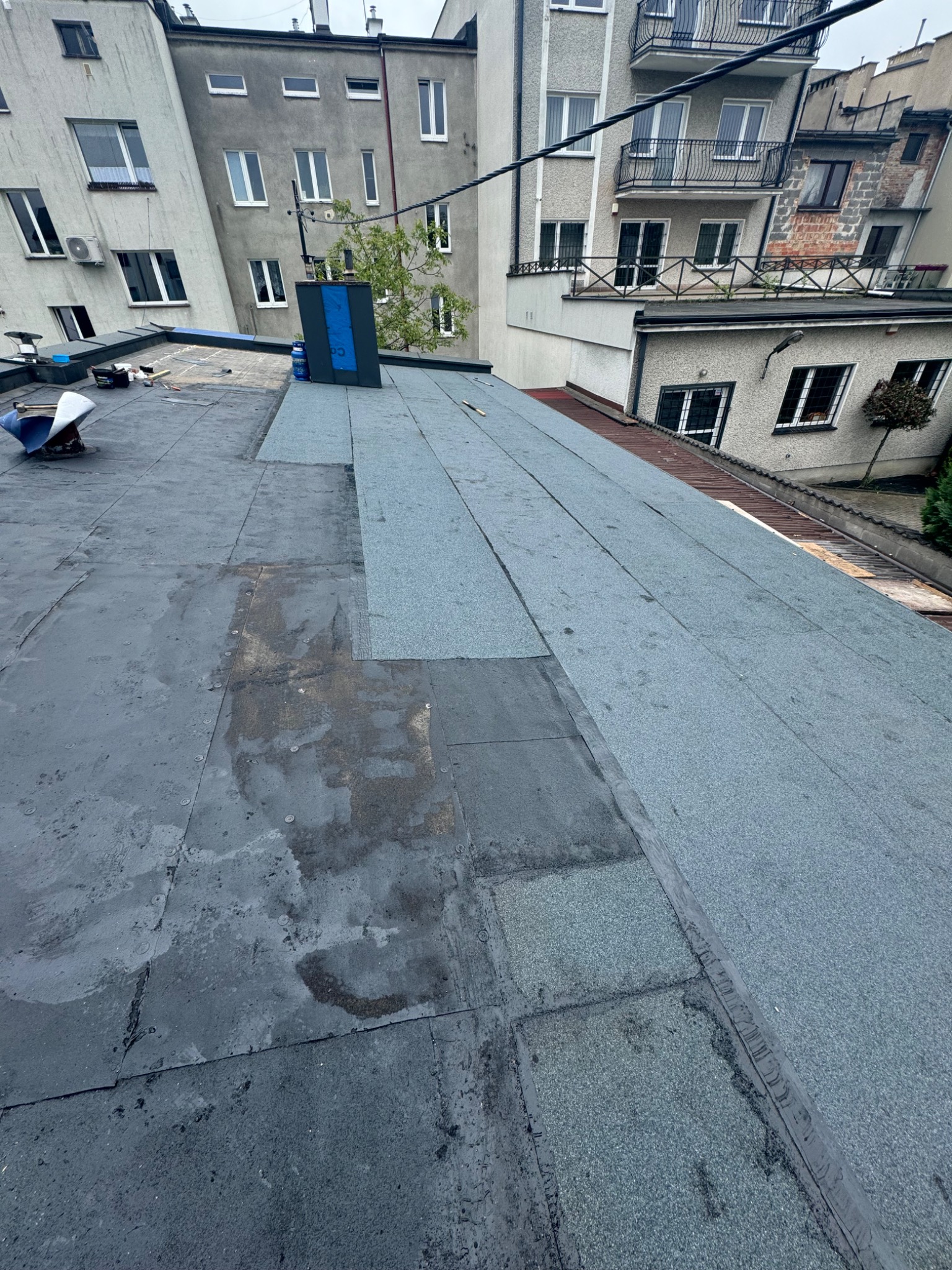 Częściowo odnowiony dach z papy na tle budynków mieszkalnych. Widoczne narzędzia i materiały dekarskie. Etap prac: wymiana starej papy na nową.