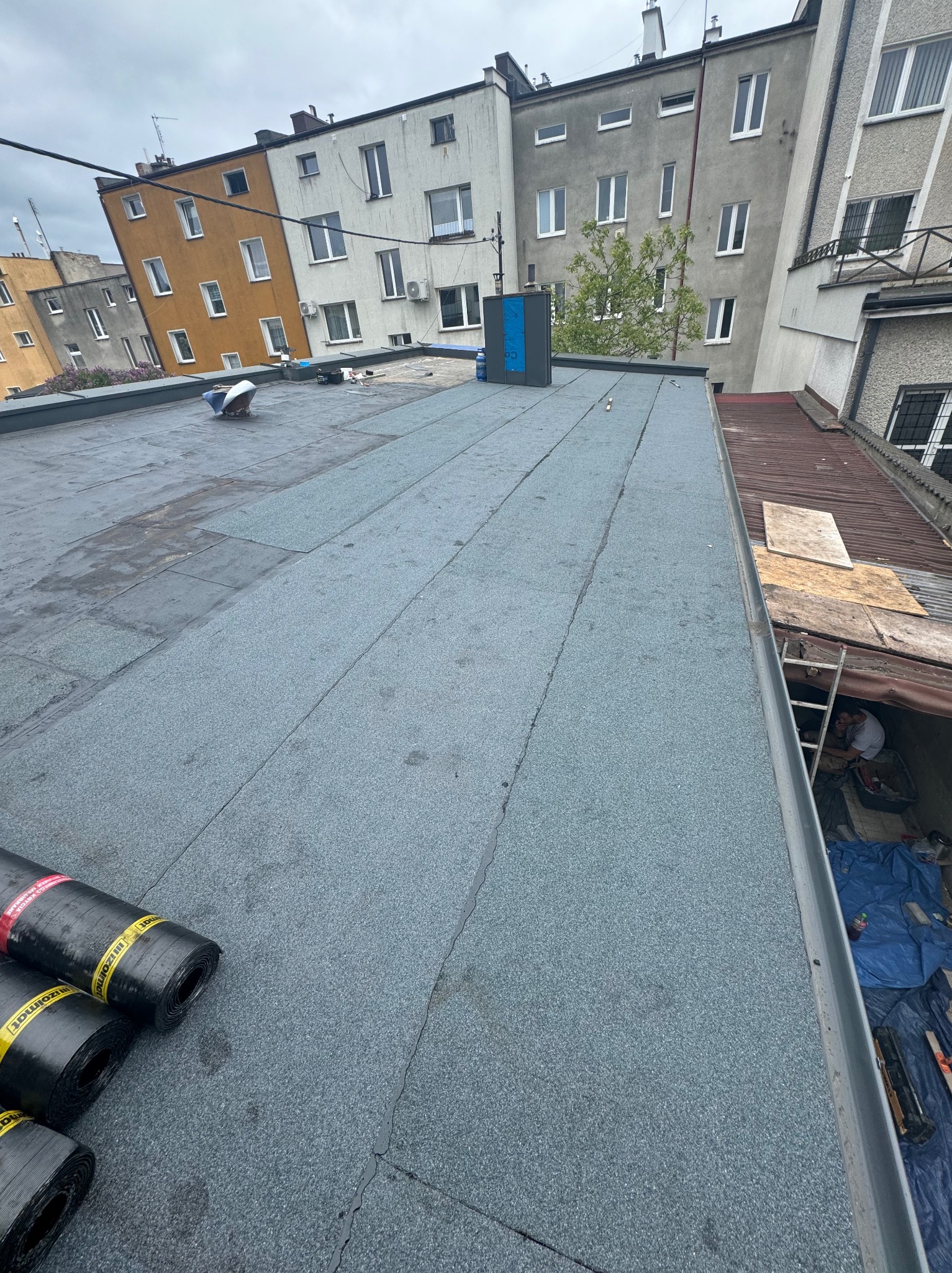 Wykończony fragment dachu pokryty papą, widoczne rolki papy na pierwszym planie, w tle budynki mieszkalne i pracownik przy krawędzi dachu.