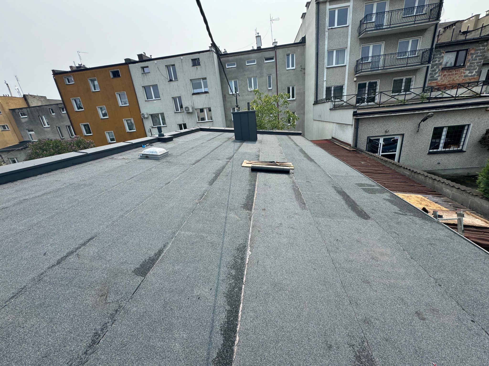 Pokrycie dachu papą termozgrzewalną na tle miejskiej zabudowy. Widoczne kominy wentylacyjne i narzędzia dekarskie. Dach po renowacji, z drobnymi śladami wilgoci.