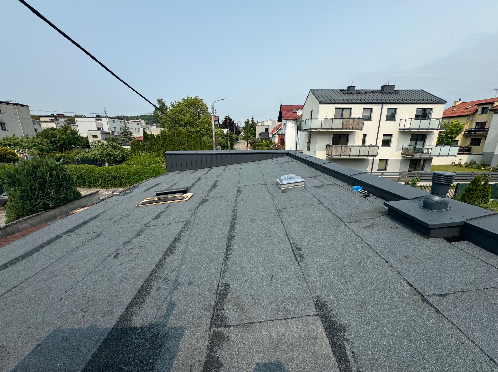 Szary dach kryty papą termozgrzewalną z świetlikami dachowymi i kominem wentylacyjnym na tle zabudowy miejskiej w słoneczny dzień. Widok z góry.