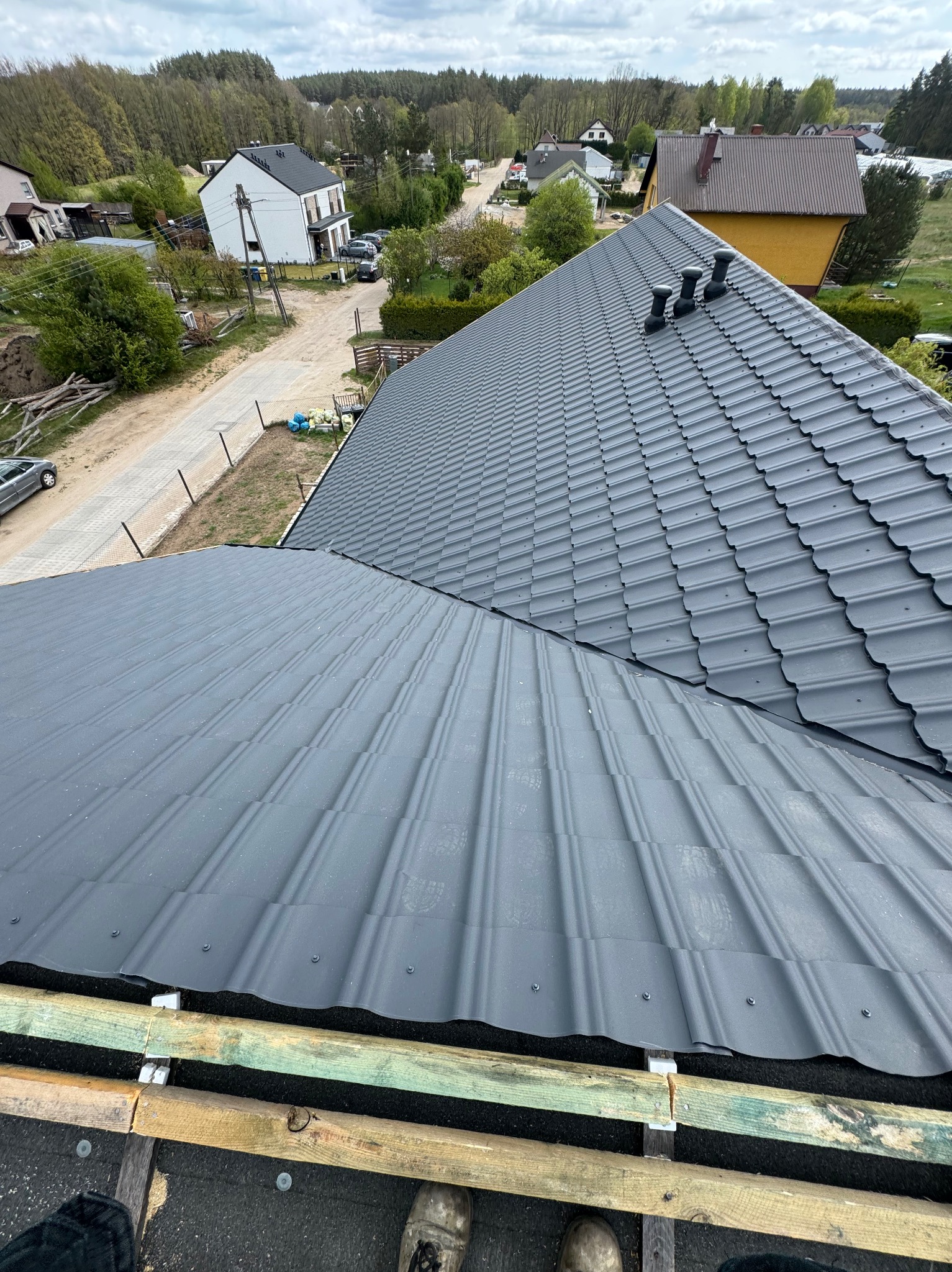 Szary dach z blachy trapezowej w trakcie montażu, widok z góry na konstrukcję więźby i elementy mocujące. W tle zabudowa jednorodzinna i zieleń.