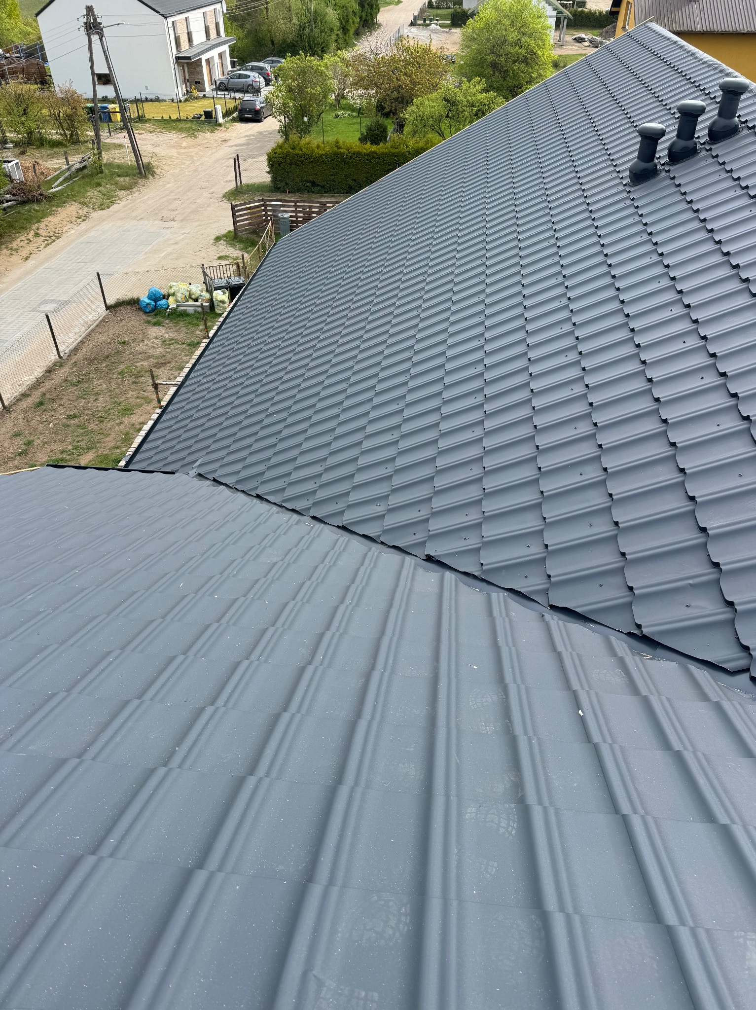 Szary dach z blachy trapezowej z trzema kominkami wentylacyjnymi, widok z góry na tle zieleni i zabudowań. Widoczne ślady po śniegu.