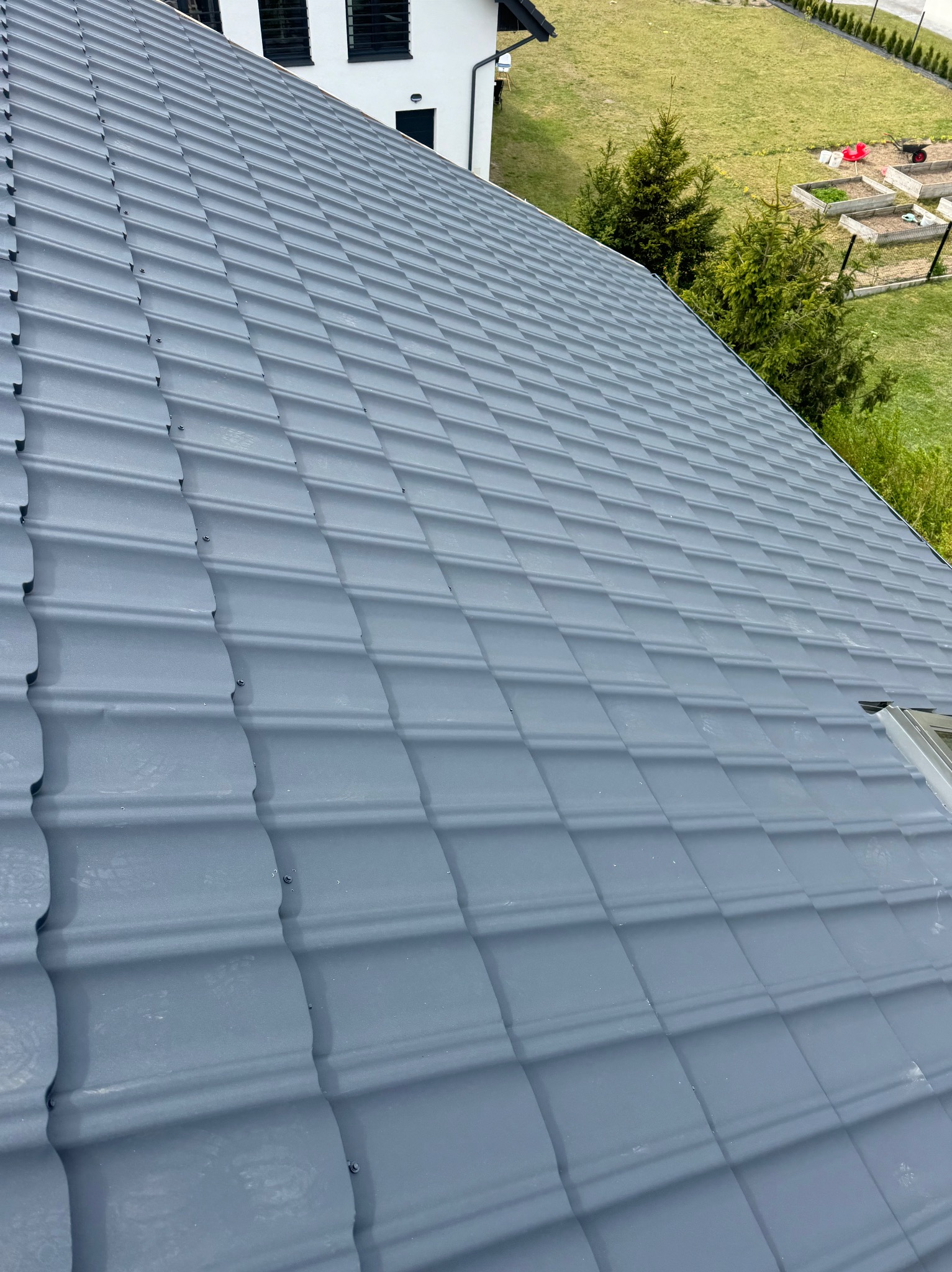 Szary dach z dachówki ceramicznej, widok z góry, fragment budynku z oknami dachowymi, w tle zieleń ogrodu. Dachówka ułożona równo, widoczne mocowania.