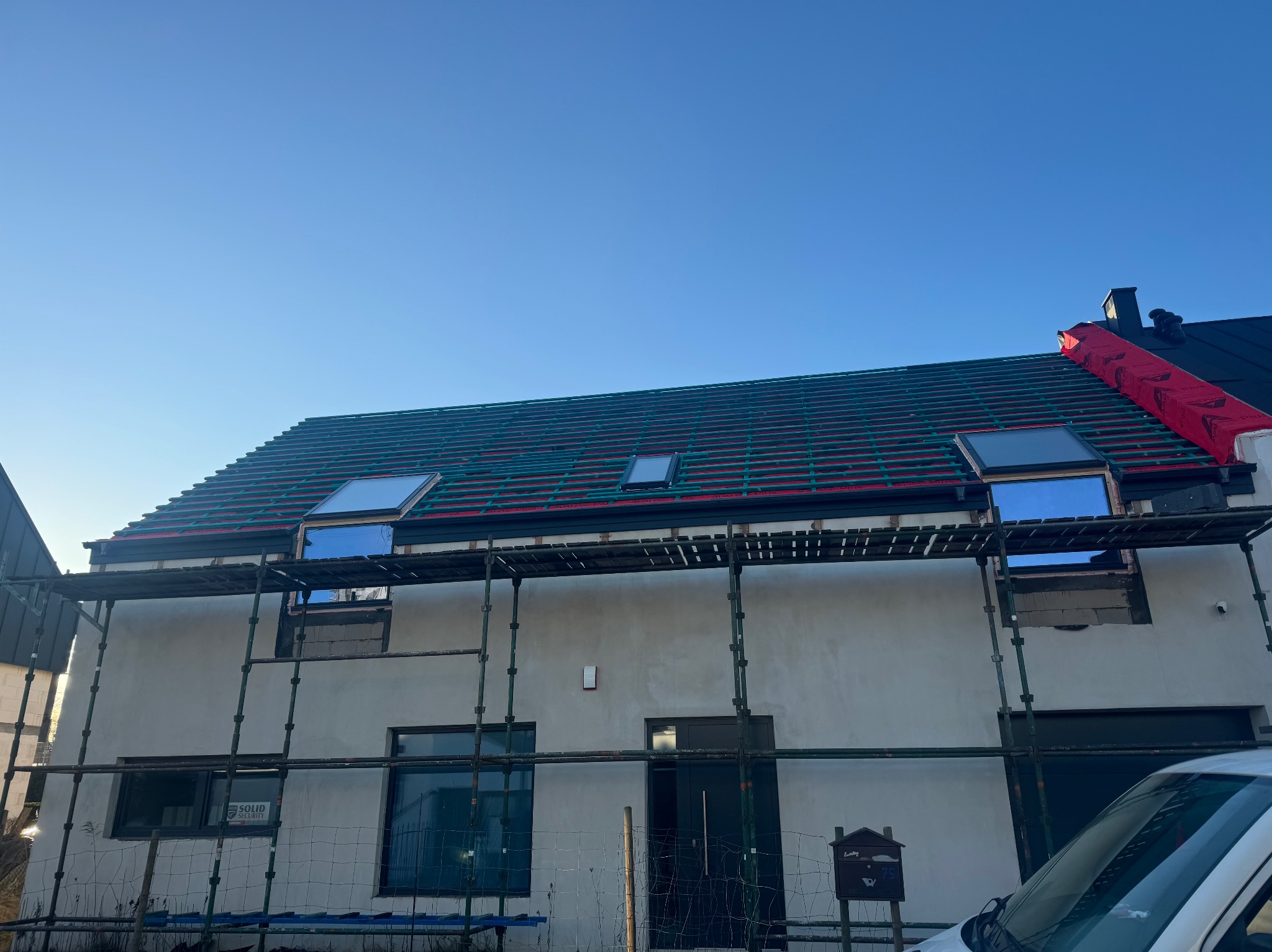 Dom w trakcie krycia dachu z oknami dachowymi, widoczne rusztowania i folia dachowa. Częściowo otynkowana elewacja, błękitne niebo w tle.