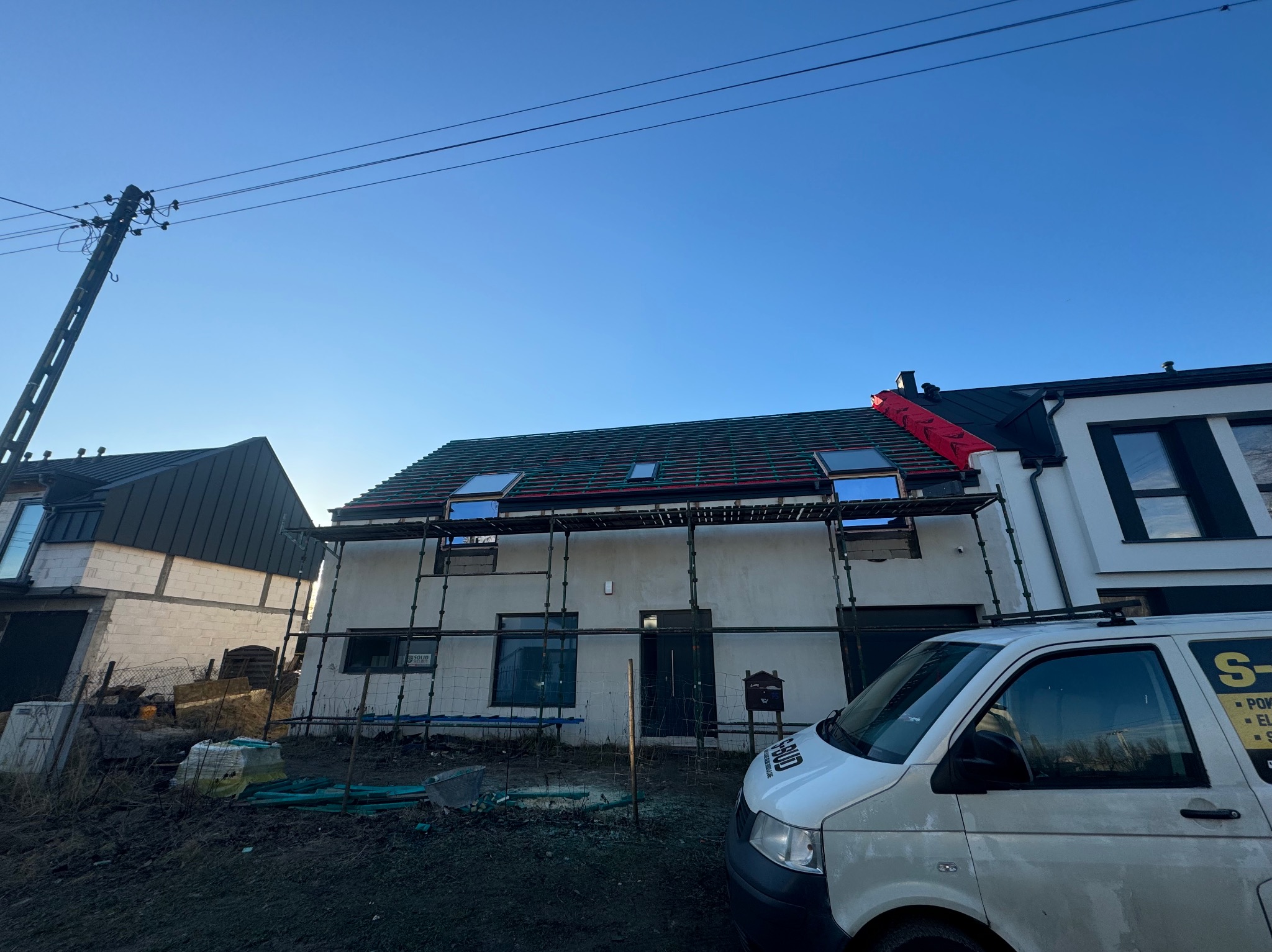 Dom w trakcie krycia dachu z oknami dachowymi, widoczne rusztowania i fragment firmowego busa. Częściowo położona dachówka, błękitne niebo w tle.