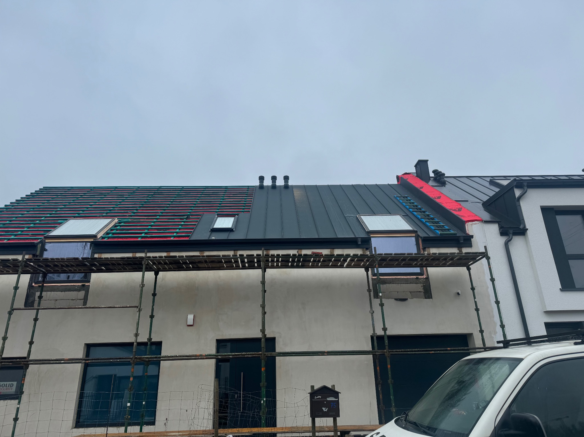 Dom w trakcie krycia dachu blachą na rąbek stojący, widoczne rusztowania i okna dachowe. Część dachu pokryta, część w trakcie prac.