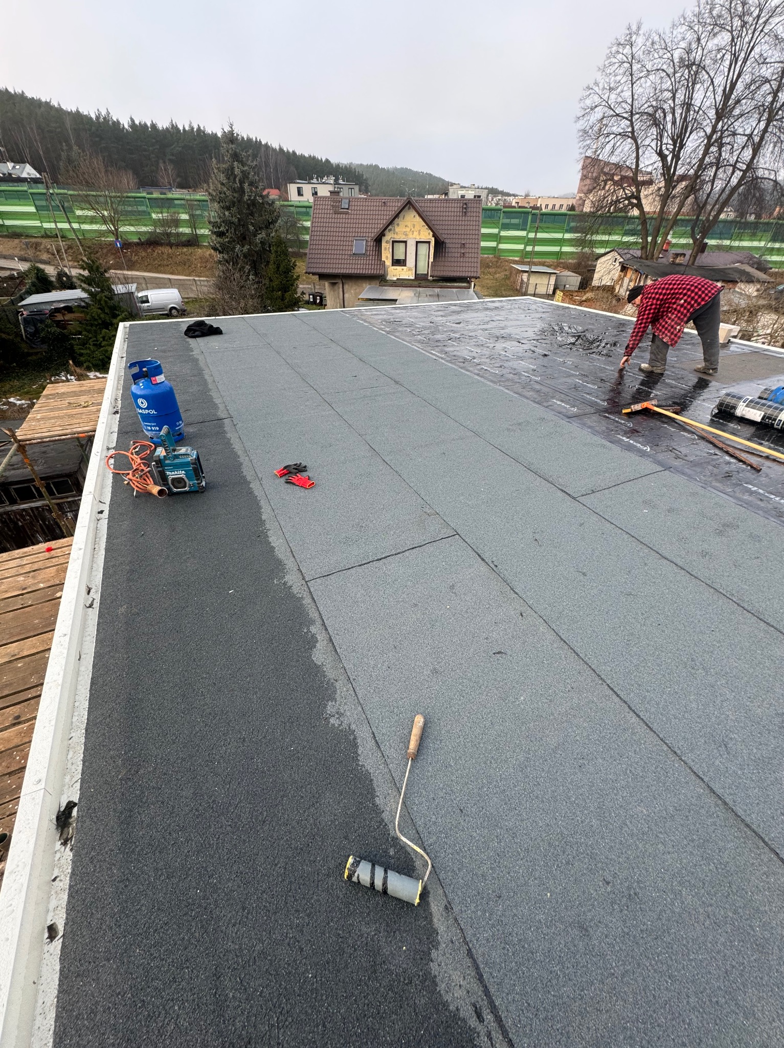 Prace dekarskie na płaskim dachu: dekarz w czerwonej koszuli układa papę termozgrzewalną. Widoczny sprzęt, butla z gazem i narzędzia dekarskie. W tle zabudowa jednorodzinna.