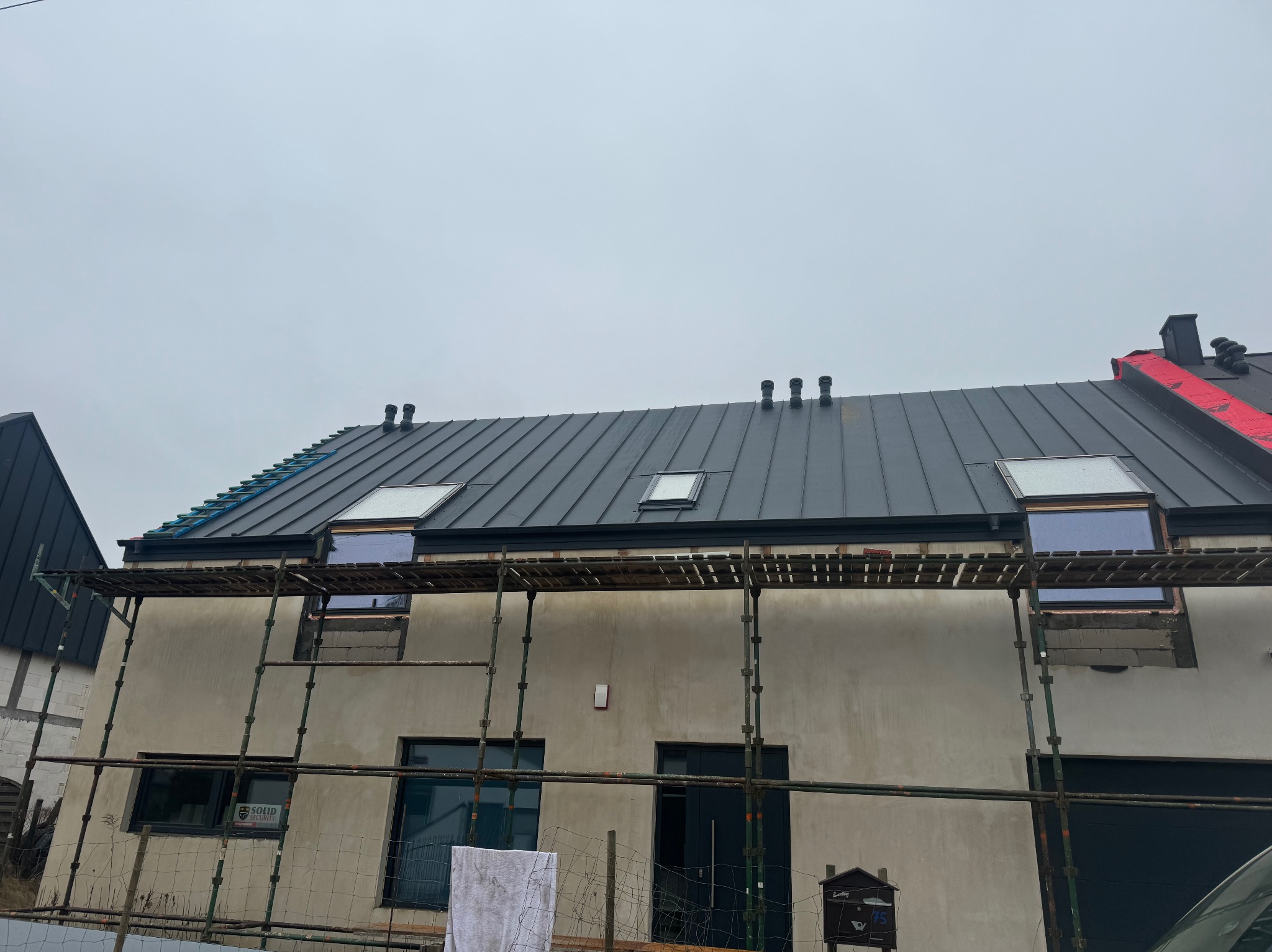 Dom w trakcie prac elewacyjnych i dekarskich: szare krycie dachu z oknami, rusztowanie, otynkowana elewacja, fragment przodu auta. Pochmurne niebo.