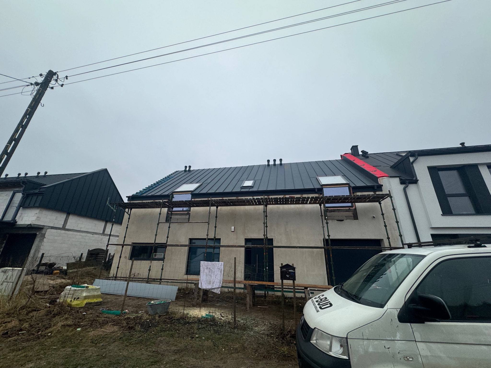 Dom w trakcie prac elewacyjnych i dekarskich, widoczne rusztowania, częściowo położony dach z oknami dachowymi oraz samochód firmy budowlanej.