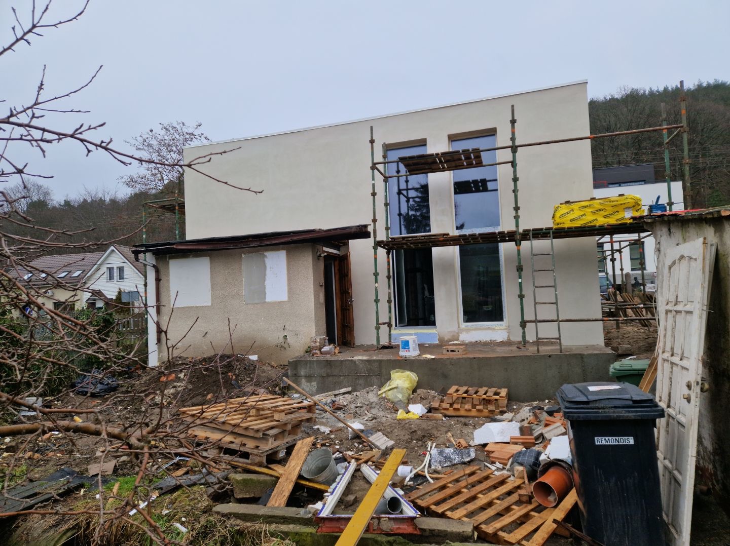 Dom w trakcie ocieplania elewacji, widoczne rusztowania, śmieci i materiały budowlane. Fragment budynku z nową elewacją. Tło: zalesione wzgórze.