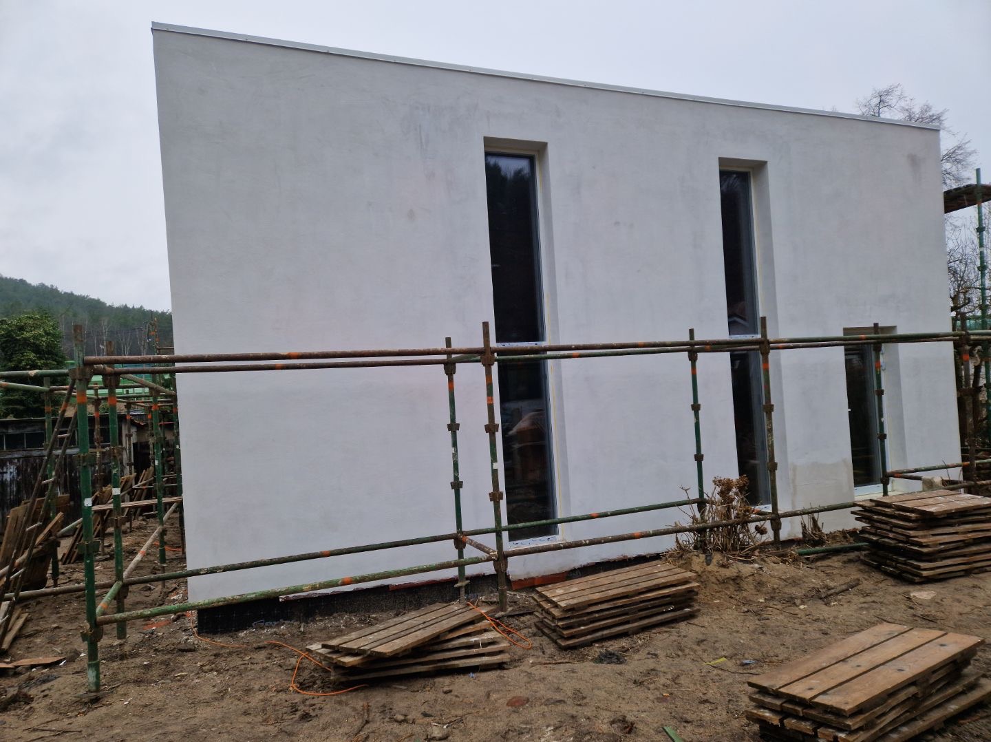 Elewacja minimalistycznego domu z dwoma pionowymi oknami, widoczna ze strony rusztowania. Tynk w jasnym odcieniu szarości, w tle zalesione wzgórze.