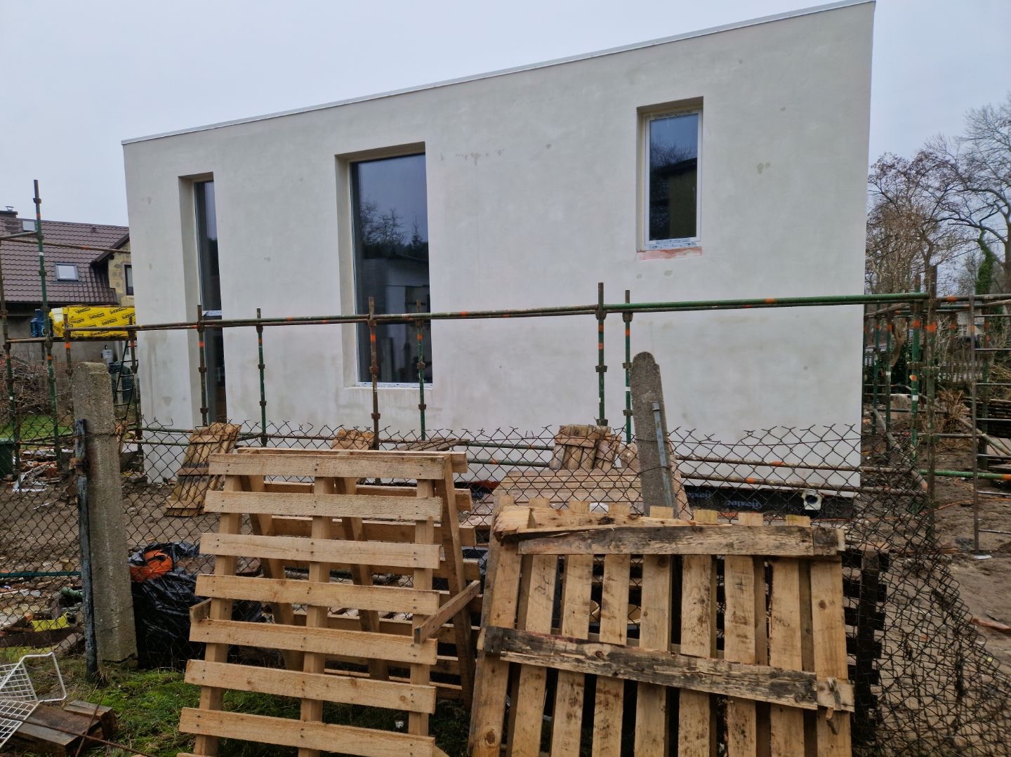 Surowa elewacja budynku z trzema oknami, widoczna siatka ogrodzeniowa i palety. Świeżo nałożony tynk elewacyjny w jasnym odcieniu szarości. Budowa w toku.