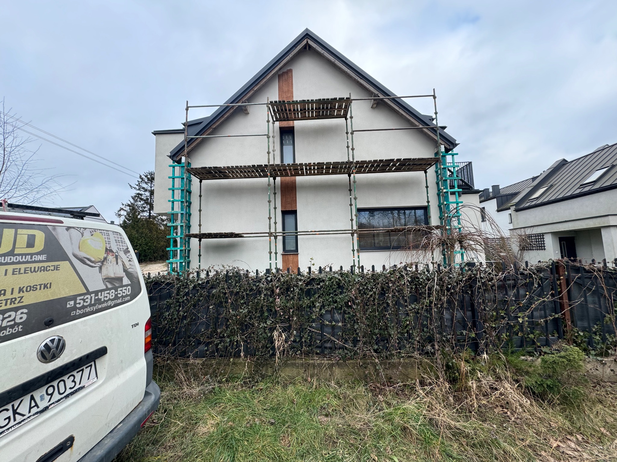 Dom z elewacją w trakcie prac, widoczne rusztowania. Na pierwszym planie fragment samochodu z reklamą firmy budowlanej. Szara, pochmurna pogoda.