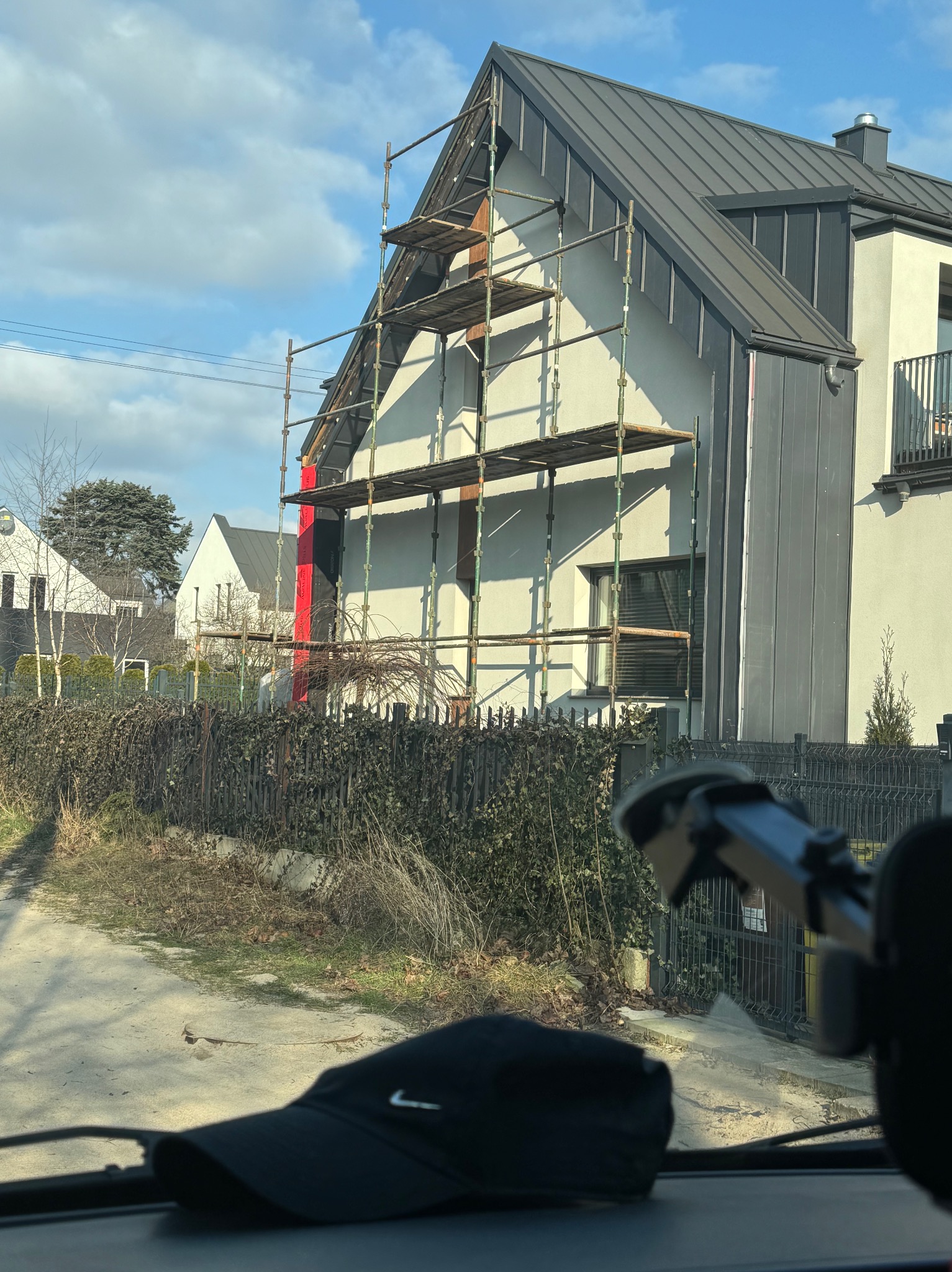 Nowoczesny dom z szarą blachą na dachu, częściowo osłonięty rusztowaniem. Fragment czerwonej izolacji na elewacji. Widok z wnętrza pojazdu.