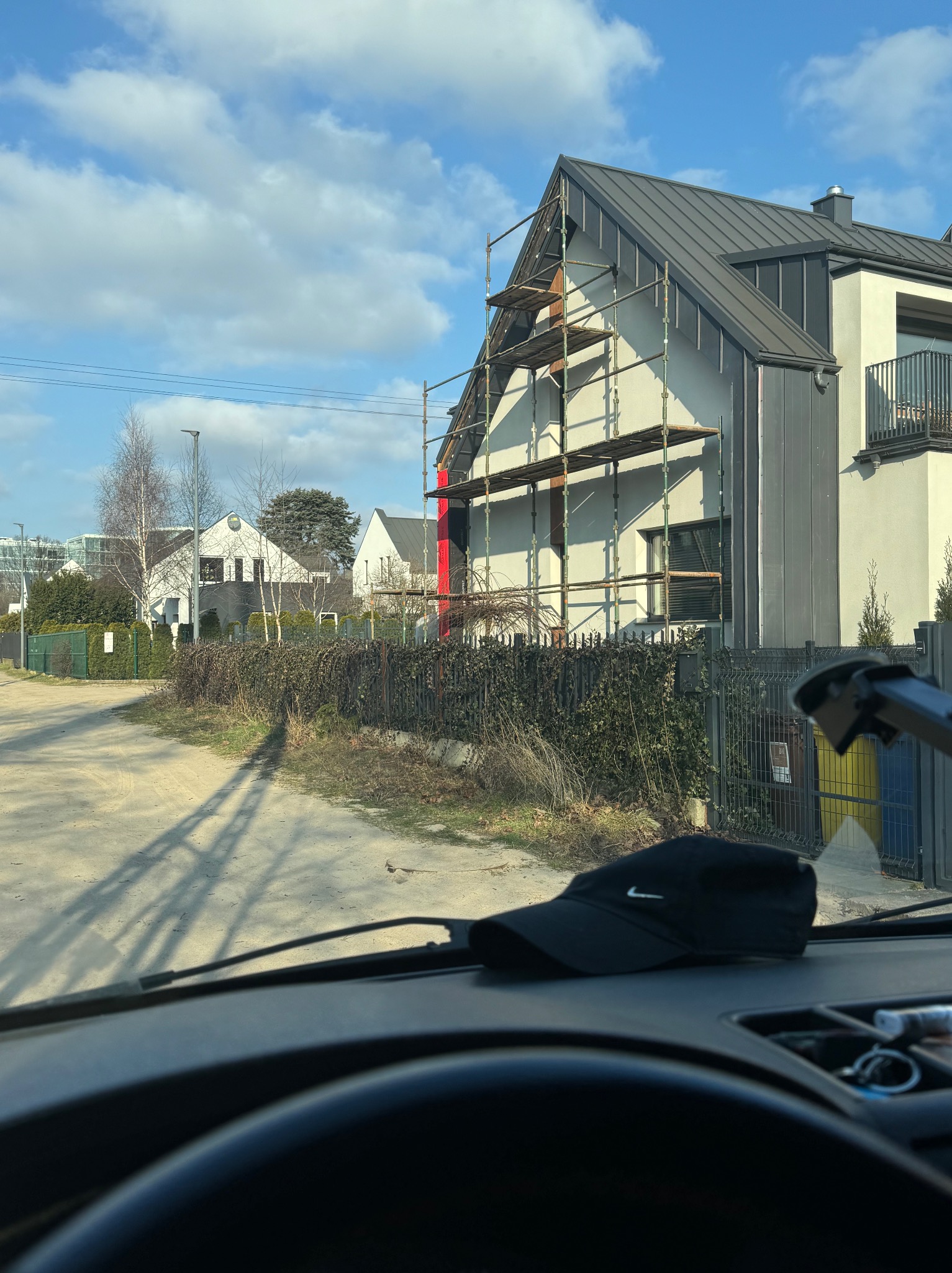 Dom w trakcie prac elewacyjnych z rusztowaniem. Widok zza szyby pojazdu, w tle inne budynki i błękitne niebo z chmurami. Czerwony element dekoracyjny na elewacji.