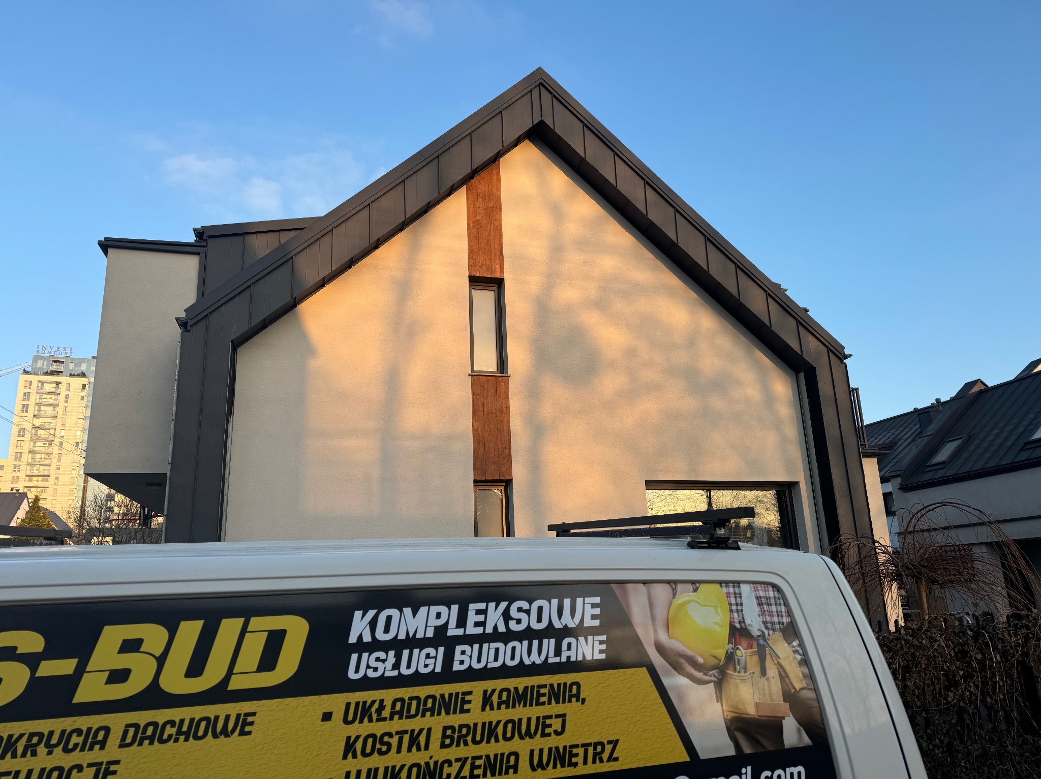 Nowoczesny dom jednorodzinny z ciemnym dachem, oświetlony popołudniowym słońcem, z fragmentem samochodu z reklamą usług budowlanych w kadrze.