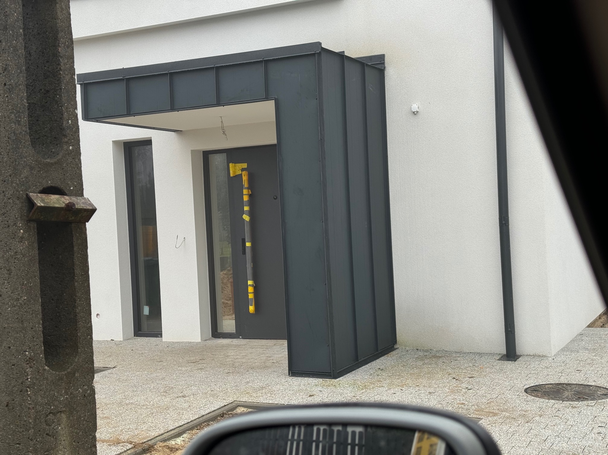 Nowoczesny dom w Gniewinie z elewacją w jasnym kolorze i ciemnymi obróbkami blacharskimi przy wejściu, widok z perspektywy przejeżdżającego samochodu.