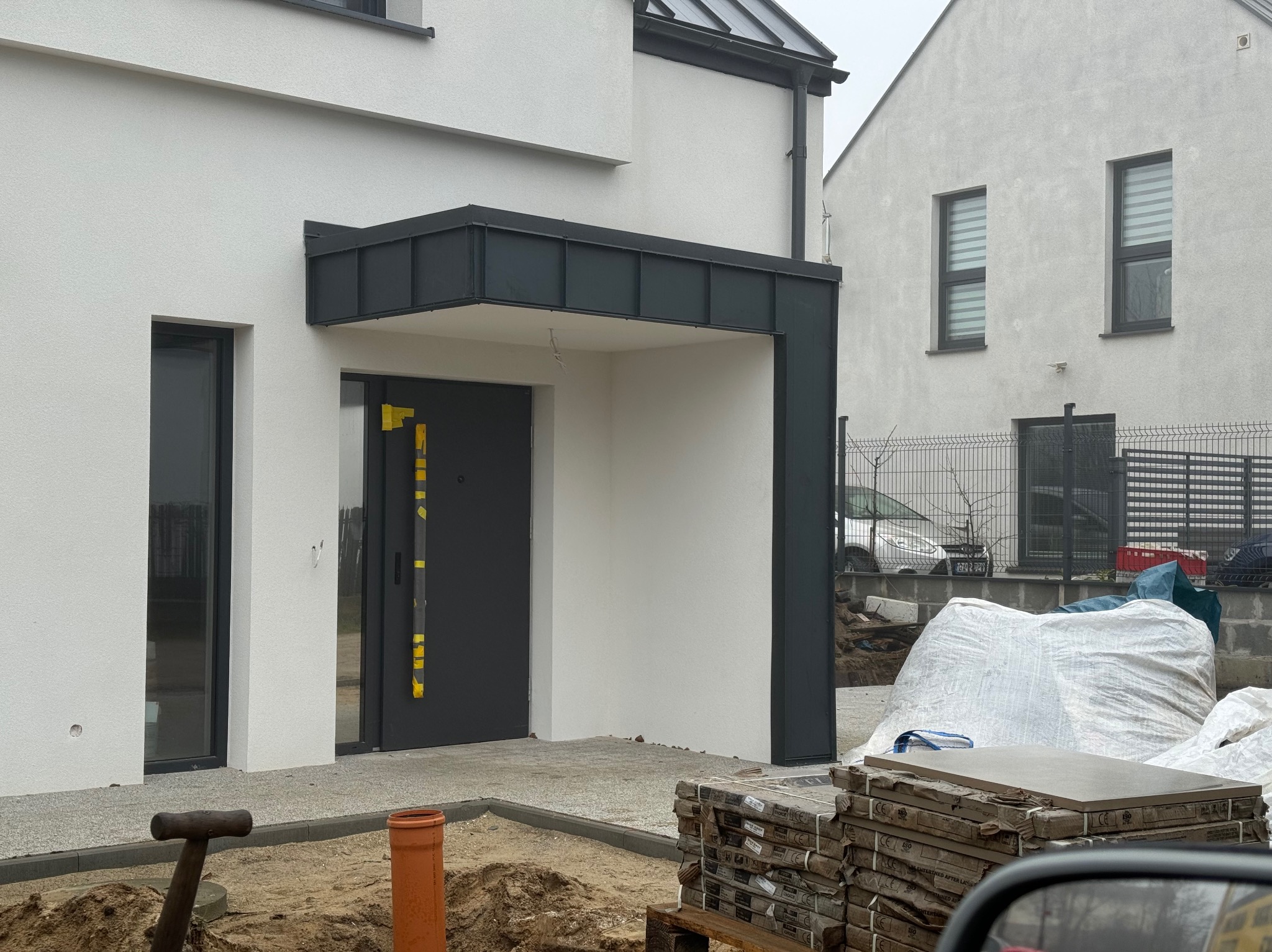 Nowoczesny dom z szarą elewacją i ciemną stolarką okienną. Widoczny fragment wejścia z zadaszeniem. Na pierwszym planie materiały budowlane i narzędzia.