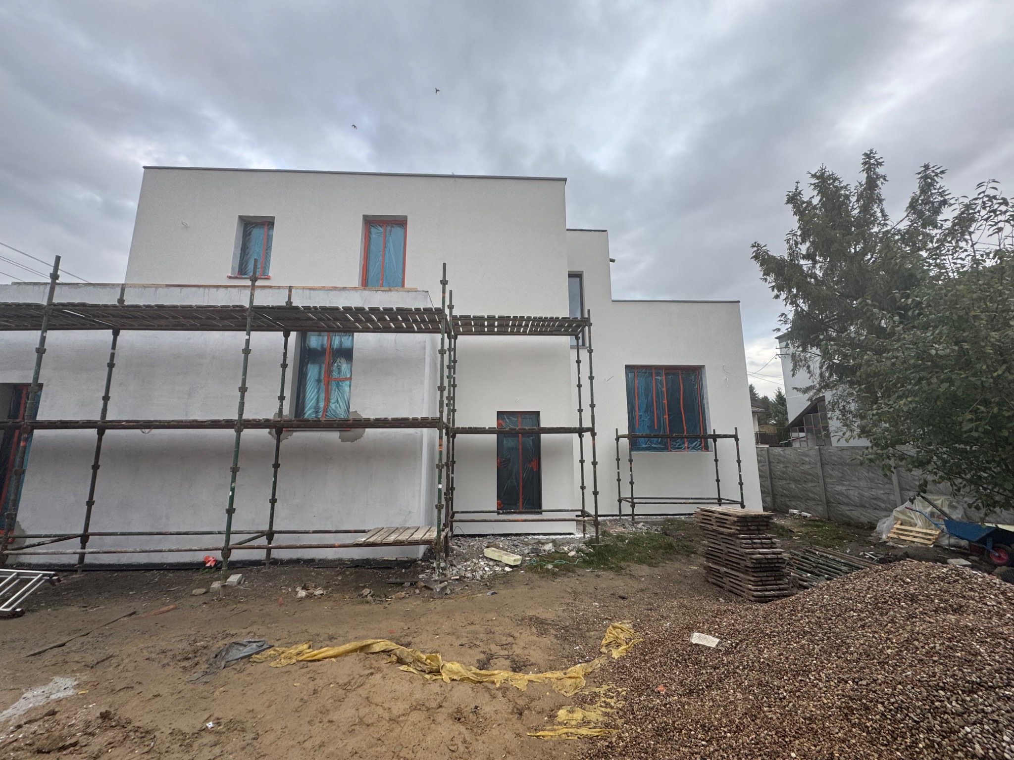 Nowoczesny dom z białą elewacją w trakcie prac wykończeniowych, z rusztowaniem, zabezpieczonymi oknami i stertą żwiru na pierwszym planie.