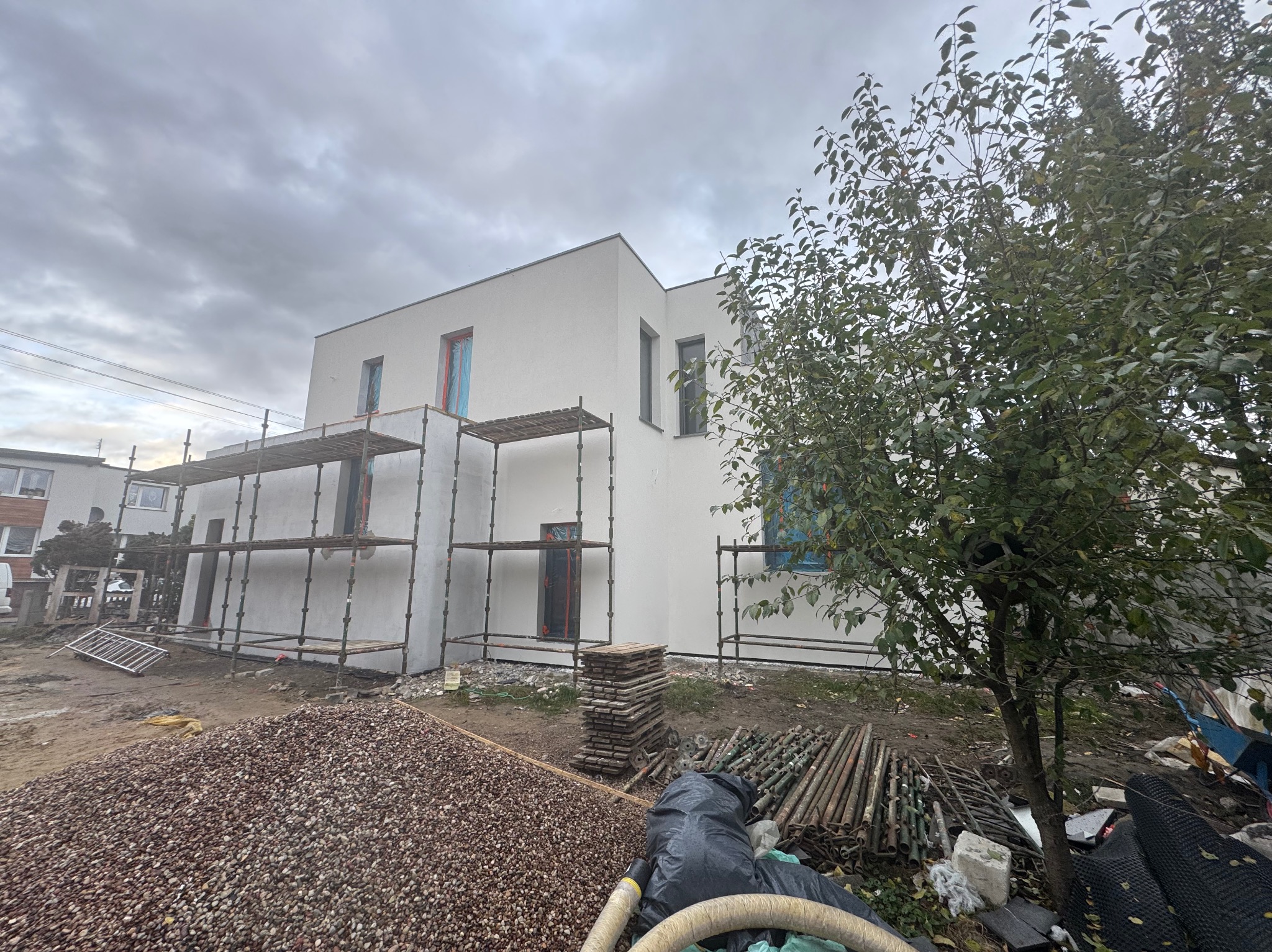 Nowoczesny, biały dom z elewacją w trakcie prac, osłonięte okna folią, rusztowania, sterta kamieni i drzewo na pierwszym planie. Prace wykończeniowe.