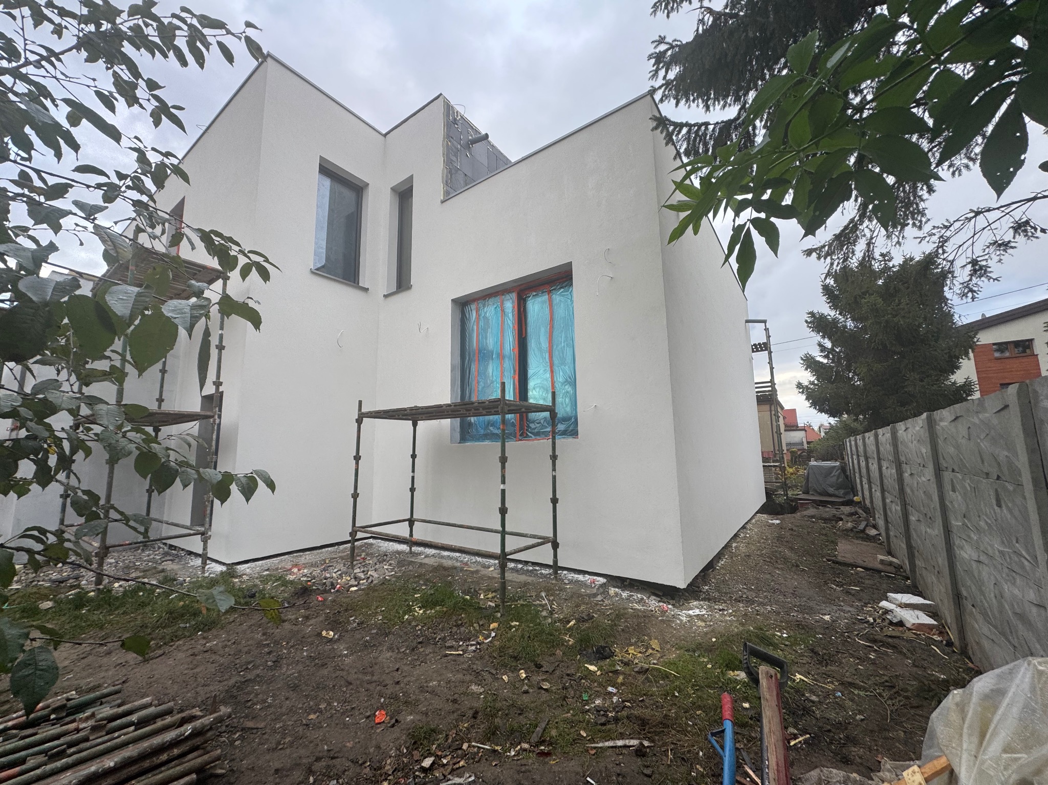 Biała elewacja nowoczesnego domu w trakcie wykańczania, okna zabezpieczone folią, widoczne rusztowania i teren budowy. Minimalistyczna architektura.