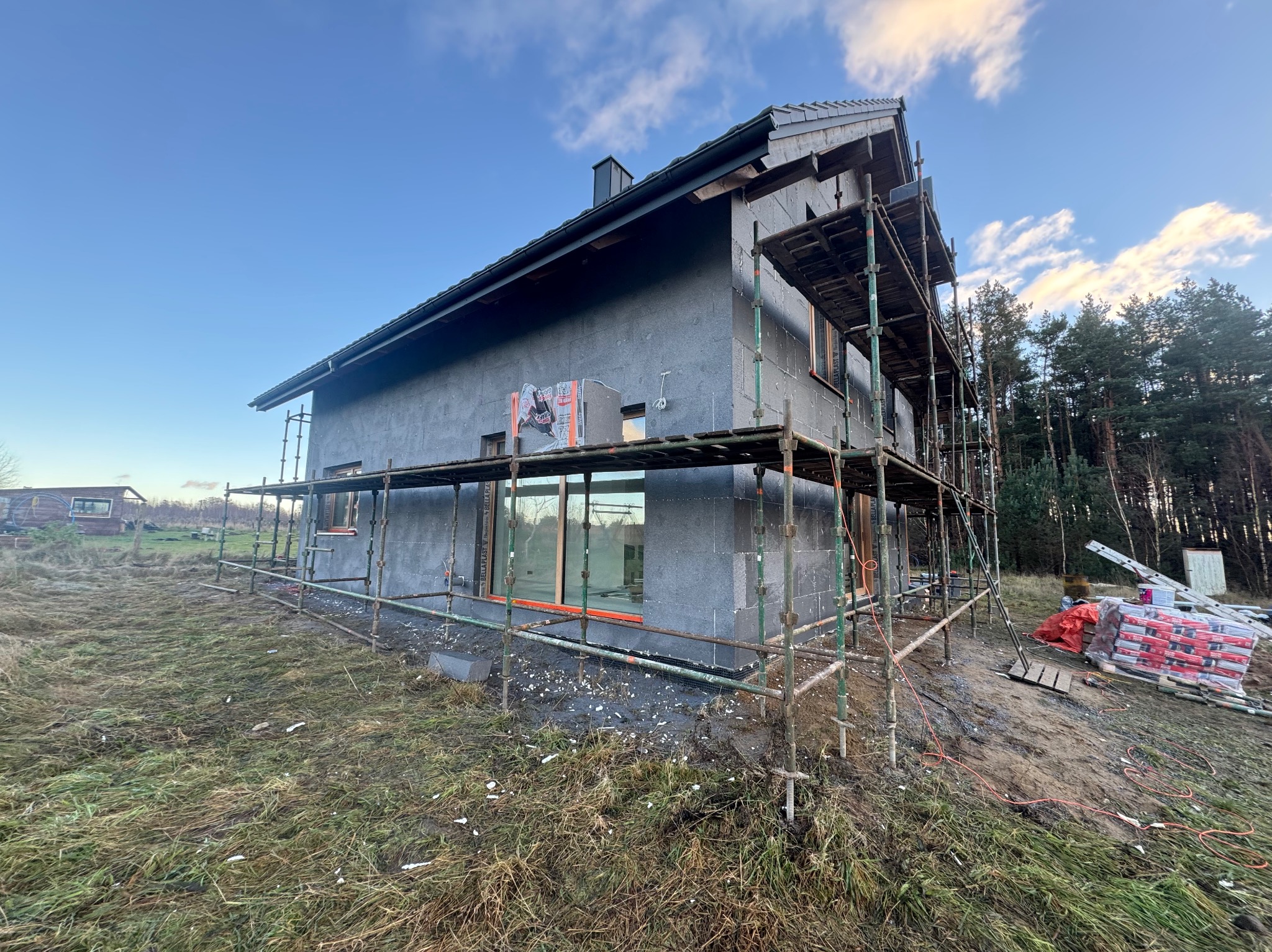 Dom w trakcie ocieplania, widoczne rusztowania i materiały izolacyjne. Szara elewacja, okna z pomarańczowymi ramami. Tło: las i błękitne niebo.