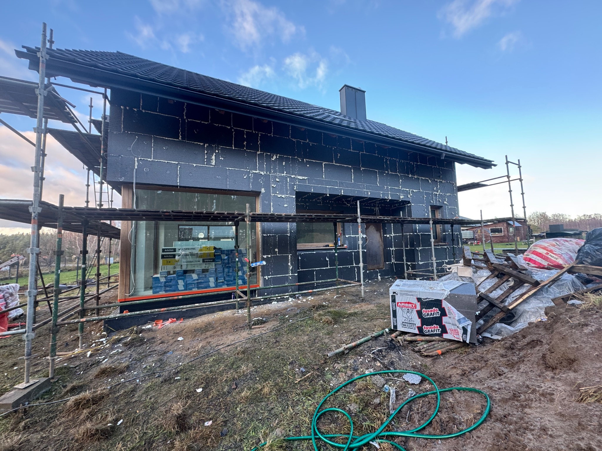 Dom w trakcie ocieplania elewacji styropianem grafitowym, widoczne rusztowania i materiały budowlane. Etap prac przed nałożeniem tynku.